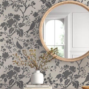 May include: A gray and white floral stencil with birds on a beige background. The stencil is being used to decorate a wall with a round mirror and a vase of branches.