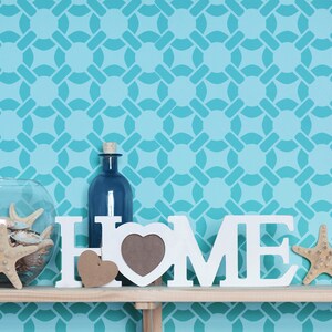 May include: A shelf displays home decor items against a turquoise patterned wall. A glass bowl with seashells, a blue bottle, and a white "HOME" sign with a heart-shaped frame are visible. Starfish and a wooden heart add to the coastal theme.