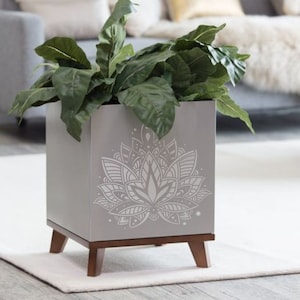 May include: A gray planter with a decorative mandala design features a leafy green plant. The planter has wooden legs.