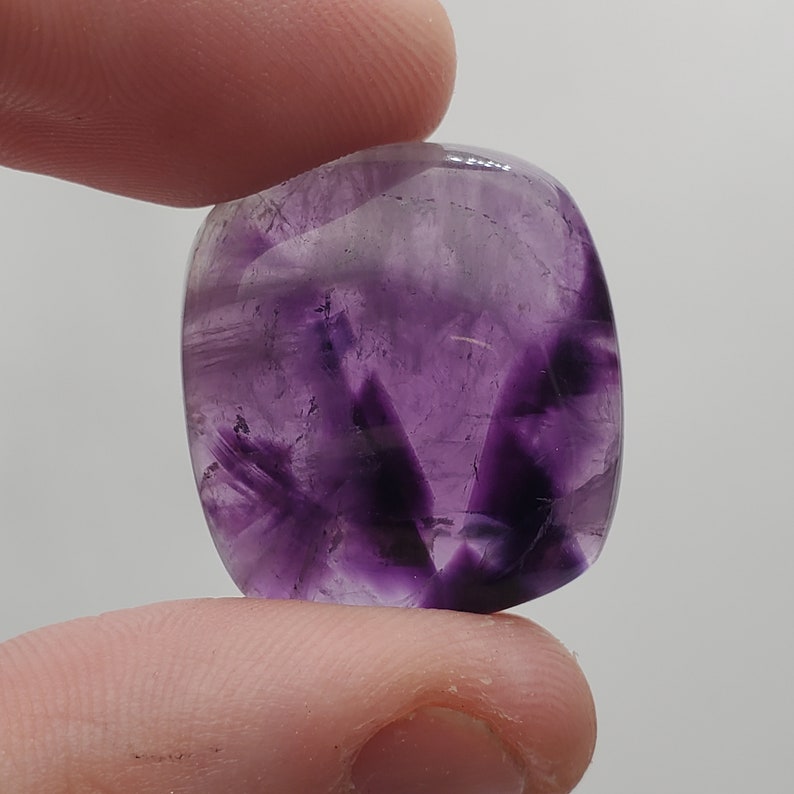 May include: A purple amethyst cabochon with a smooth, polished surface. The stone has a swirling pattern of light and dark purple hues.