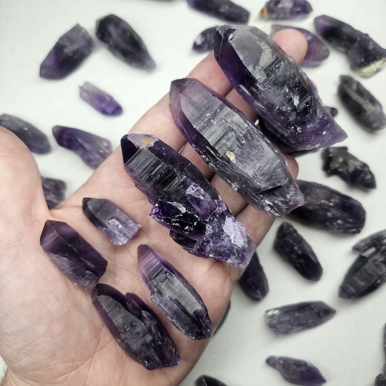Peut inclure: Plusieurs cristaux d'am&eacute;thyste bruts de tailles et de formes vari&eacute;es. Les cristaux pr&eacute;sentent des nuances de violet, du fonc&eacute; au clair, avec des facettes visibles et des textures naturelles. Certains cristaux sont tenus dans une main, d'autres sont dispers&eacute;s en arri&egrave;re-plan.