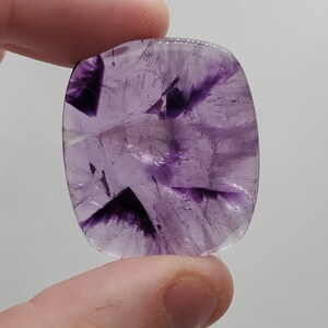 May include: A purple amethyst cabochon with a unique, swirling pattern. The stone is translucent and has a light purple hue.