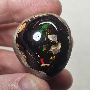 May include: A dark brown, black and green opal with a rough, natural surface. The opal has a rainbow sheen and is set in a rough, natural matrix.