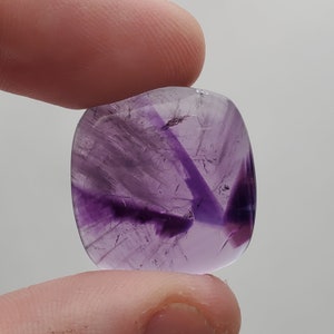 May include: A purple amethyst cabochon with a unique, swirling pattern of lighter and darker shades of purple. The stone is smooth and polished, with a slightly rounded shape.