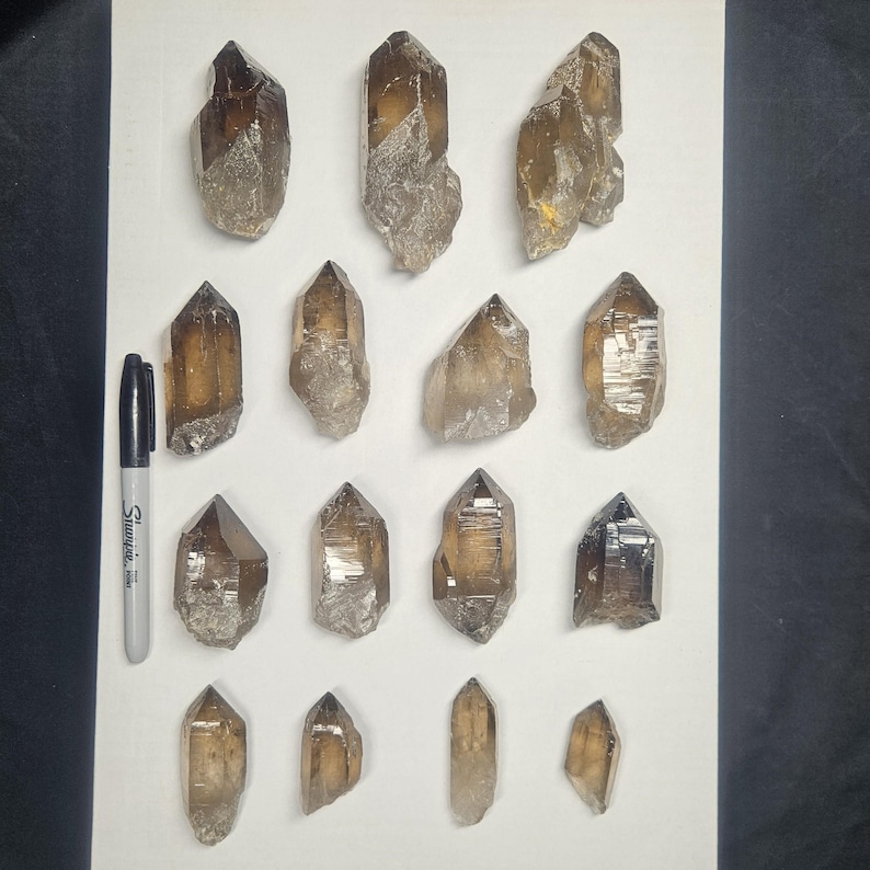 May include: A collection of smoky quartz crystal points in various sizes and shapes. The crystals range in color from light to dark brown, with some having a translucent appearance. A black Sharpie marker is included for size reference.
