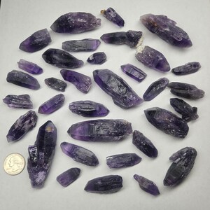 Peut inclure: Une collection de cristaux d'am&eacute;thyste bruts de formes et tailles vari&eacute;es. Les cristaux pr&eacute;sentent des nuances de violet, du fonc&eacute; au clair, avec des textures et des facettes naturelles. Une pi&egrave;ce de monnaie est incluse pour l'&eacute;chelle, mesurant les cristaux en centim&egrave;tres.