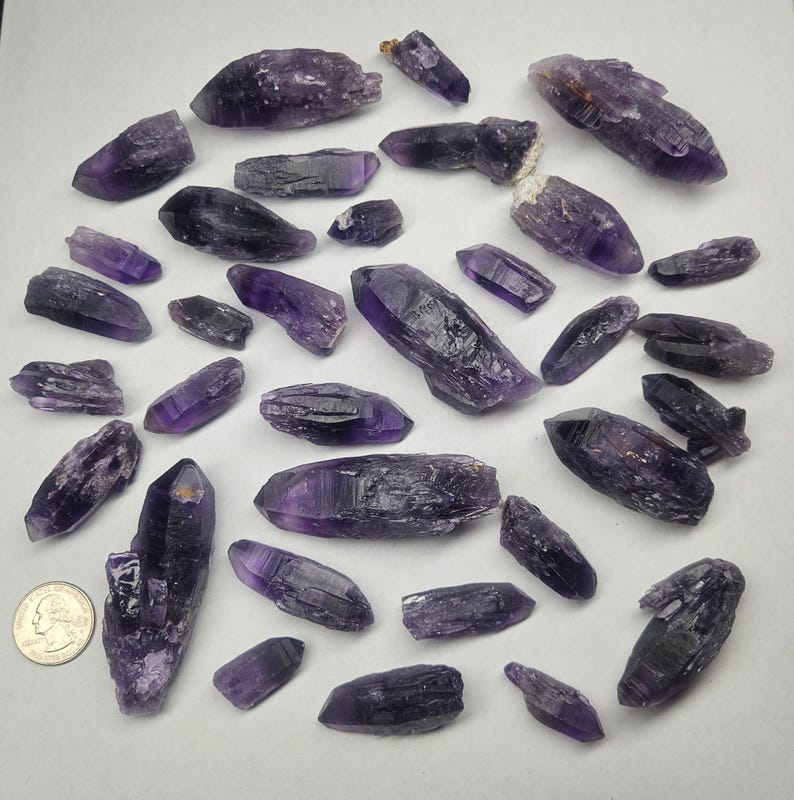 Peut inclure: Une collection de cristaux d'am&eacute;thyste bruts de formes et tailles vari&eacute;es. Les cristaux pr&eacute;sentent des nuances de violet, du fonc&eacute; au clair, avec des textures et des facettes naturelles. Une pi&egrave;ce de monnaie est incluse pour l'&eacute;chelle, mesurant les cristaux en centim&egrave;tres.
