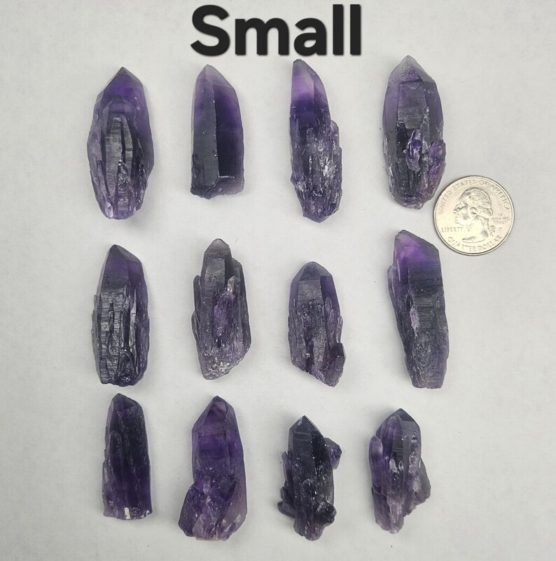 Peut inclure: Plusieurs petits cristaux d'am&eacute;thyste bruts de diff&eacute;rentes nuances de violet. Les cristaux sont dispos&eacute;s sur une surface blanche, avec une pi&egrave;ce de monnaie pour l'&eacute;chelle. Le mot "Small" est imprim&eacute; en noir en haut.