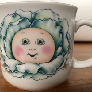 May include: White ceramic mug featuring a cartoon illustration of a baby's face inside a green cabbage.