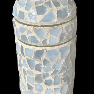 Peut inclure: Un vase en céramique blanche avec un motif en mosaïque composé de morceaux de verre bleu clair. Le vase a une forme cylindrique et un sommet légèrement évasé.