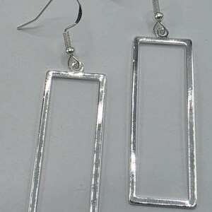 May include: A pair of silver-toned earrings featuring a simple, open rectangular frame design. Each earring hangs from a standard curved ear wire hook, showcasing a polished metallic finish that reflects light across the smooth, thin rectangular edges.
