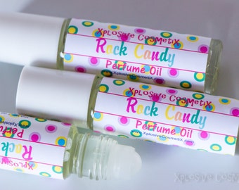 May include: Three clear glass roll-on perfume oil bottles with white caps. Each bottle has a label with the text "Rock Candy Perfume Oil" and a colorful polka dot design. The brand name "Xplosive Cosmetix" is also visible.