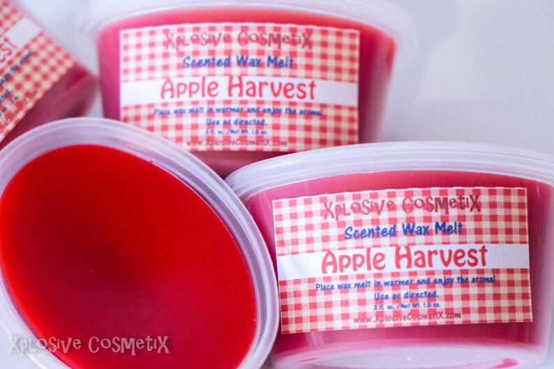 May include: Three red wax melts in clear plastic containers with white lids. Each container has a red and white checkered label that reads "XPLOSIVE COSMETIX Scented Wax Melt Apple Harvest".
