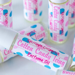 K&ouml;nnte beinhalten: Mehrere kleine Glasflaschen mit Parf&uuml;m&ouml;l und wei&szlig;en Kappen liegen auf einer Oberfl&auml;che. Die Etiketten zeigen rosa und blaue Tupfen und den Text "Cotton Candy Icing Perfume Oil" in Rosa und Blau. Der Markenname "Xplosive Cosmetix" ist ebenfalls zu sehen.