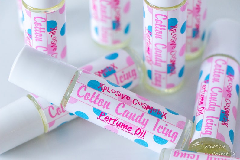 K&ouml;nnte beinhalten: Mehrere kleine Glasflaschen mit Parf&uuml;m&ouml;l und wei&szlig;en Kappen liegen auf einer Oberfl&auml;che. Die Etiketten zeigen rosa und blaue Tupfen und den Text "Cotton Candy Icing Perfume Oil" in Rosa und Blau. Der Markenname "Xplosive Cosmetix" ist ebenfalls zu sehen.