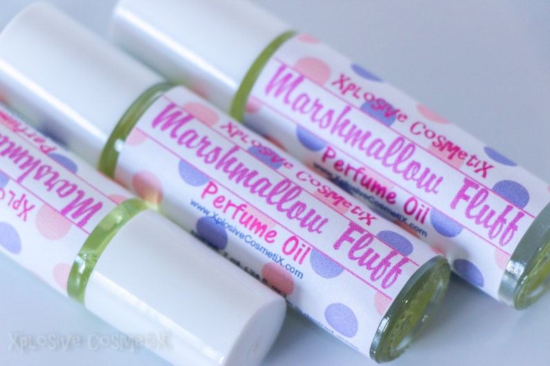 May include: Several perfume oil bottles are displayed, each with a white label featuring pink and purple polka dots. The labels read "Marshmallow Fluff Perfume Oil" in pink script, with the brand name "Xplosive CosmetiX". The bottles have white caps.