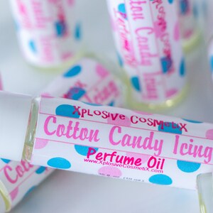 K&ouml;nnte beinhalten: Mehrere Parf&uuml;m&ouml;lflaschen mit wei&szlig;en Kappen und Etiketten. Die Etiketten zeigen "Cotton Candy Icing Perfume Oil" in Rosa und Blau, mit dem Markennamen "Xplosive Cosmetix". Die Flaschen sind mit einer klaren Fl&uuml;ssigkeit gef&uuml;llt.