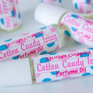 K&ouml;nnte beinhalten: Mehrere Parf&uuml;m&ouml;lflaschen mit wei&szlig;en Kappen liegen auf einer wei&szlig;en Oberfl&auml;che. Die Etiketten tragen die Aufschrift "Cotton Candy Icing Perfume Oil" mit rosa und blauen Tupfen. Der Markenname "Xplosive Cosmetix" ist ebenfalls sichtbar.