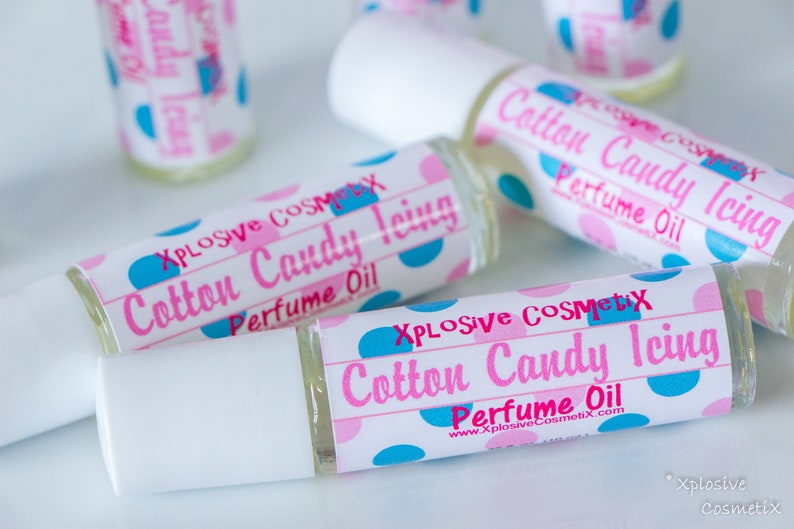 K&ouml;nnte beinhalten: Mehrere Parf&uuml;m&ouml;lflaschen mit wei&szlig;en Kappen liegen auf einer wei&szlig;en Oberfl&auml;che. Die Etiketten tragen die Aufschrift "Cotton Candy Icing Perfume Oil" mit rosa und blauen Tupfen. Der Markenname "Xplosive Cosmetix" ist ebenfalls sichtbar.