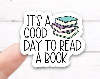 May include: White sticker with black text that reads "It's a good day to read a book". A stack of colorful books is illustrated next to the text.