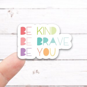 May include: A white sticker with the words "Be Kind", "Be Brave", and "Be You" in colorful letters. The sticker is being held by a finger.