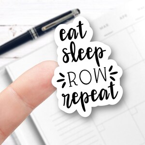 May include: White sticker with black text that reads "eat sleep row repeat". A finger is holding the sticker up against a white planner.
