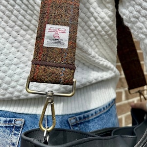 Puede incluir: Una correa de hombro de tweed marr&oacute;n y beige con un cierre de metal dorado y una etiqueta blanca que dice "Harris Tweed" con un peque&ntilde;o logotipo de un p&aacute;jaro y el texto "Handwoven" y "Outer Hebrides" en una fuente m&aacute;s peque&ntilde;a.