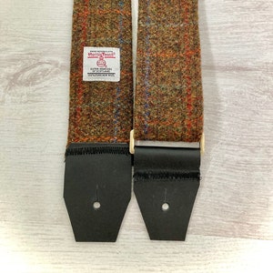 May include: Two brown tweed guitar straps with black leather ends and gold metal buckles. The strap on the left has a label that reads "Hand Woven Cloth Harris Tweed Outer Hebrides Scotland 100% Pure New Wool".