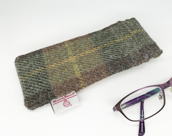 May include: A green, brown, and yellow plaid tweed glasses case with a "Harris Tweed" label. The case is lying on a white surface next to a pair of purple-framed glasses.