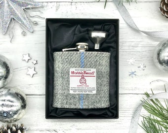 May include: A silver flask with a funnel, wrapped in gray Harris Tweed fabric with blue stripes. The flask is in a black box with a satin lining. The fabric has the Harris Tweed logo and text: "HAND WOVEN CLOTH Harris Tweed HARRIS TWEED OUTER HEBRIDES OF SCOTLAND 100% PURE NEW WOOL".