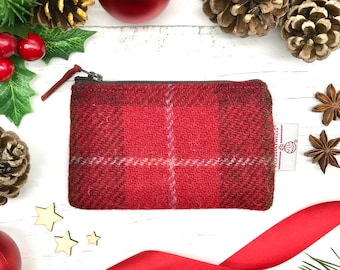 Könnte beinhalten: Rote karierte Reißverschlusstasche mit einem braunen Reißverschluss und einem kleinen Etikett mit der Aufschrift "Harris Tweed". Die Tasche liegt auf einer weißen Holzoberfläche mit rotem Band, Kiefernästen und sternförmigen Dekorationen.