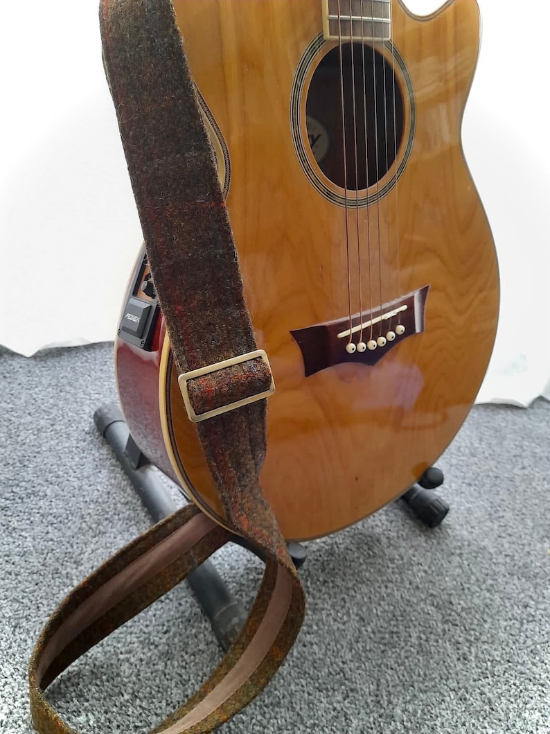 May include: A brown and tan guitar strap with a gold buckle. The strap is hanging on a wooden acoustic guitar with a sound hole and six strings.