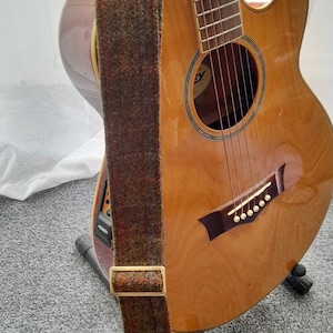 May include: A brown acoustic guitar with a tweed guitar strap. The guitar is on a stand and has a black pick-up.