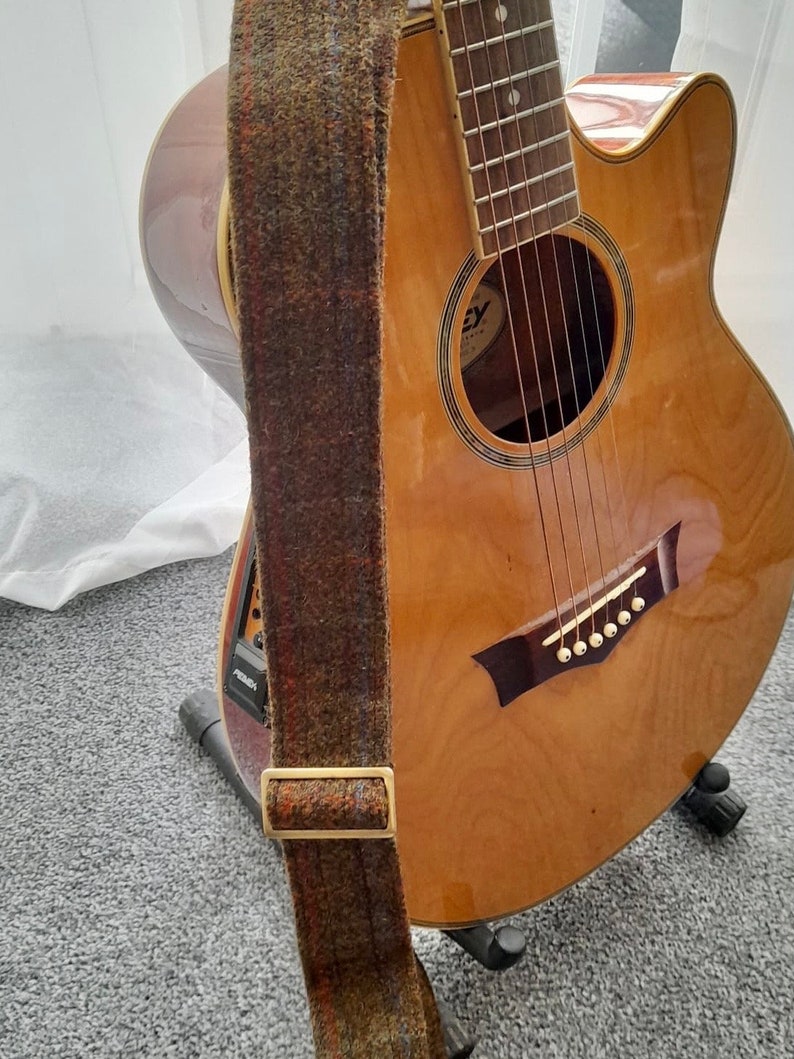 May include: A brown acoustic guitar with a tweed guitar strap. The guitar is on a stand and has a black pick-up.