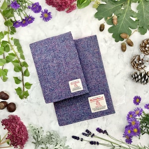 May include: Two purple and blue tweed notebook covers with a white label that says "Harris Tweed" on each. The covers are on a white surface with flowers and other natural elements around them.