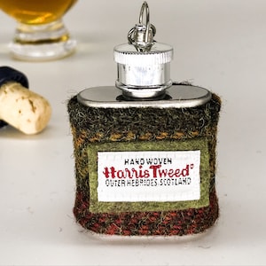 Pode incluir: Uma garrafa de prata com uma capa de tecido xadrez verde, marrom e vermelho. O tecido tem uma etiqueta que diz "Hand Woven Harris Tweed Outer Hebrides Scotland".
