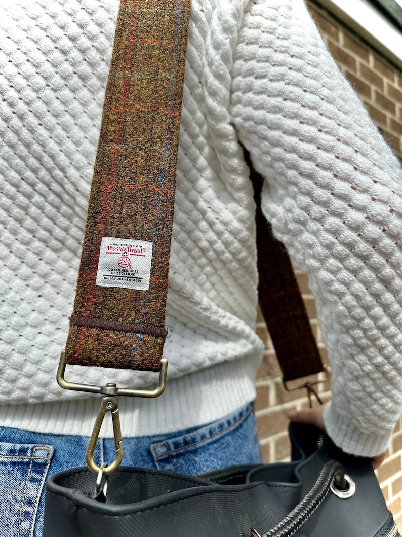 Puede incluir: Una correa de hombro de tweed marr&oacute;n y beige con un cierre de metal dorado y una etiqueta blanca que dice "Harris Tweed" con un peque&ntilde;o logotipo de un p&aacute;jaro y el texto "Handwoven" y "Outer Hebrides" en una fuente m&aacute;s peque&ntilde;a.