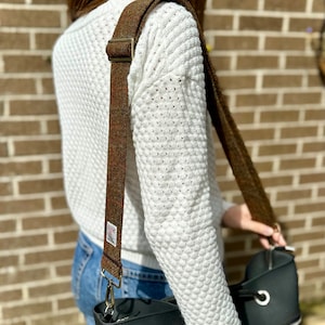 Puede incluir: Un bolso tote de cuero negro con una correa de hombro de tweed marr&oacute;n y beige. La correa tiene una hebilla dorada y un cierre plateado.