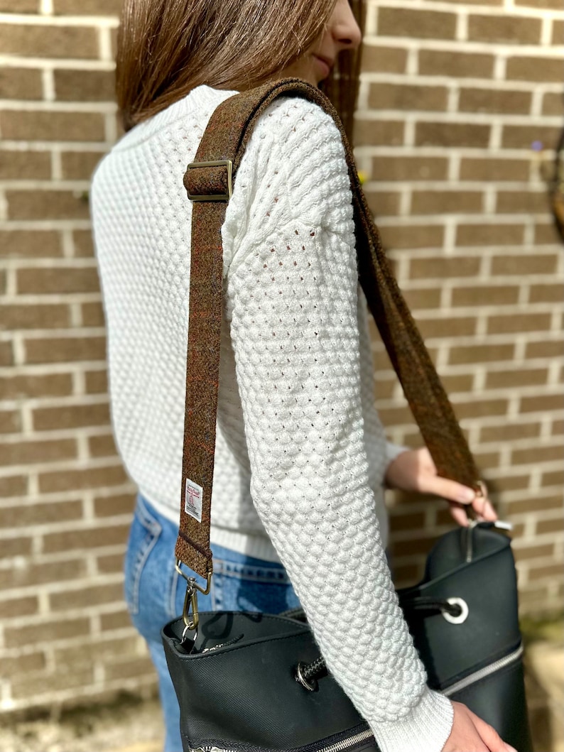 Puede incluir: Un bolso tote de cuero negro con una correa de hombro de tweed marr&oacute;n y beige. La correa tiene una hebilla dorada y un cierre plateado.