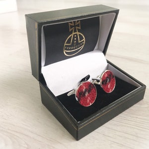 May include: A pair of cufflinks in a black box. The cufflinks are round and have a red and black tartan pattern. The box has a gold logo on the lid.