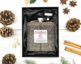May include: A stainless steel hip flask with a brown and green Harris Tweed fabric cover. The flask is in a black box with a silver funnel. The box is on a white wooden surface with pine branches, cinnamon sticks, and star anise.