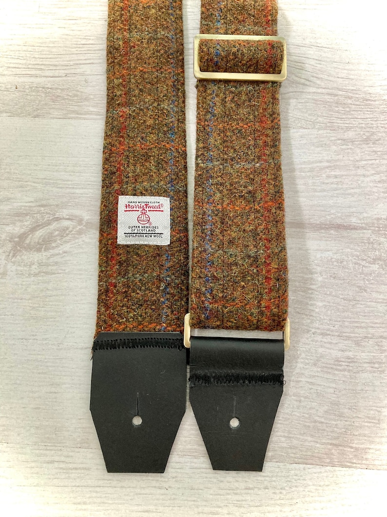 May include: Two brown tweed guitar straps with black leather ends and gold metal buckles. The strap on the left has a label that reads "Hand Woven Cloth Harris Tweed Outer Hebrides Scotland 100% Pure New Wool".