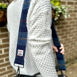 Puede incluir: Un su&eacute;ter blanco con una correa de hombro de cuadros azules y rojos unida a un bolso negro. La correa tiene una peque&ntilde;a etiqueta con el texto "Harris Tweed".
