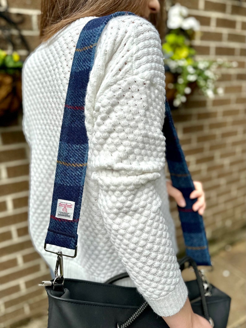 Puede incluir: Un su&eacute;ter blanco con una correa de hombro de cuadros azules y rojos unida a un bolso negro. La correa tiene una peque&ntilde;a etiqueta con el texto "Harris Tweed".