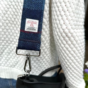 Puede incluir: Un bolso tote negro con una correa de cuadros azules y blancos. La correa tiene una etiqueta que dice "Hand Woven Cloth Harris Tweed Outer Hebrides Scotland 100% Pure New Wool".
