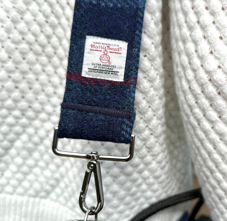Puede incluir: Una correa de tela azul y roja a cuadros con una etiqueta blanca que dice "HAND WOVEN CLOTH Harris Tweed OUTER HEBRIDES OF SCOTLAND 100% PURE NEW WOOL". La correa tiene un gancho de metal plateado.