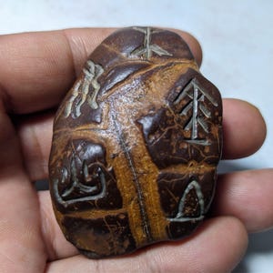 May include: A brown, irregularly shaped stone with etched symbols. The stone has a textured surface and a mix of brown and tan colours. The symbols appear to be ancient or stylized, with a tree-like design and other geometric shapes.