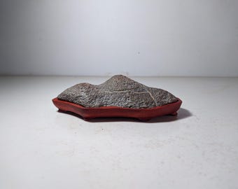 May include: A gray stone shaped like a mountain sits on a red wooden base. The stone has a rough texture and is a popular choice for bonsai displays.
