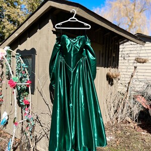 Puede incluir: Un vestido largo de sat&eacute;n verde esmeralda con escote de coraz&oacute;n y corpi&ntilde;o ajustado. El vestido tiene una falda completa y est&aacute; colgado en una percha blanca.