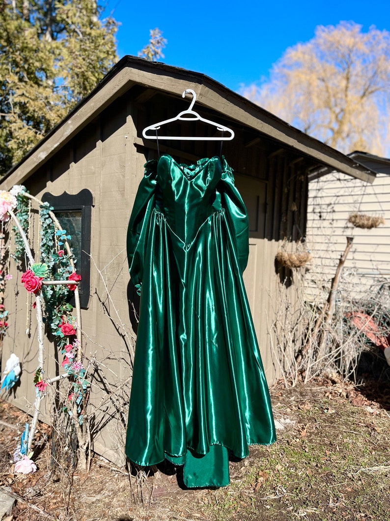 Puede incluir: Un vestido largo de sat&eacute;n verde esmeralda con escote de coraz&oacute;n y corpi&ntilde;o ajustado. El vestido tiene una falda completa y est&aacute; colgado en una percha blanca.